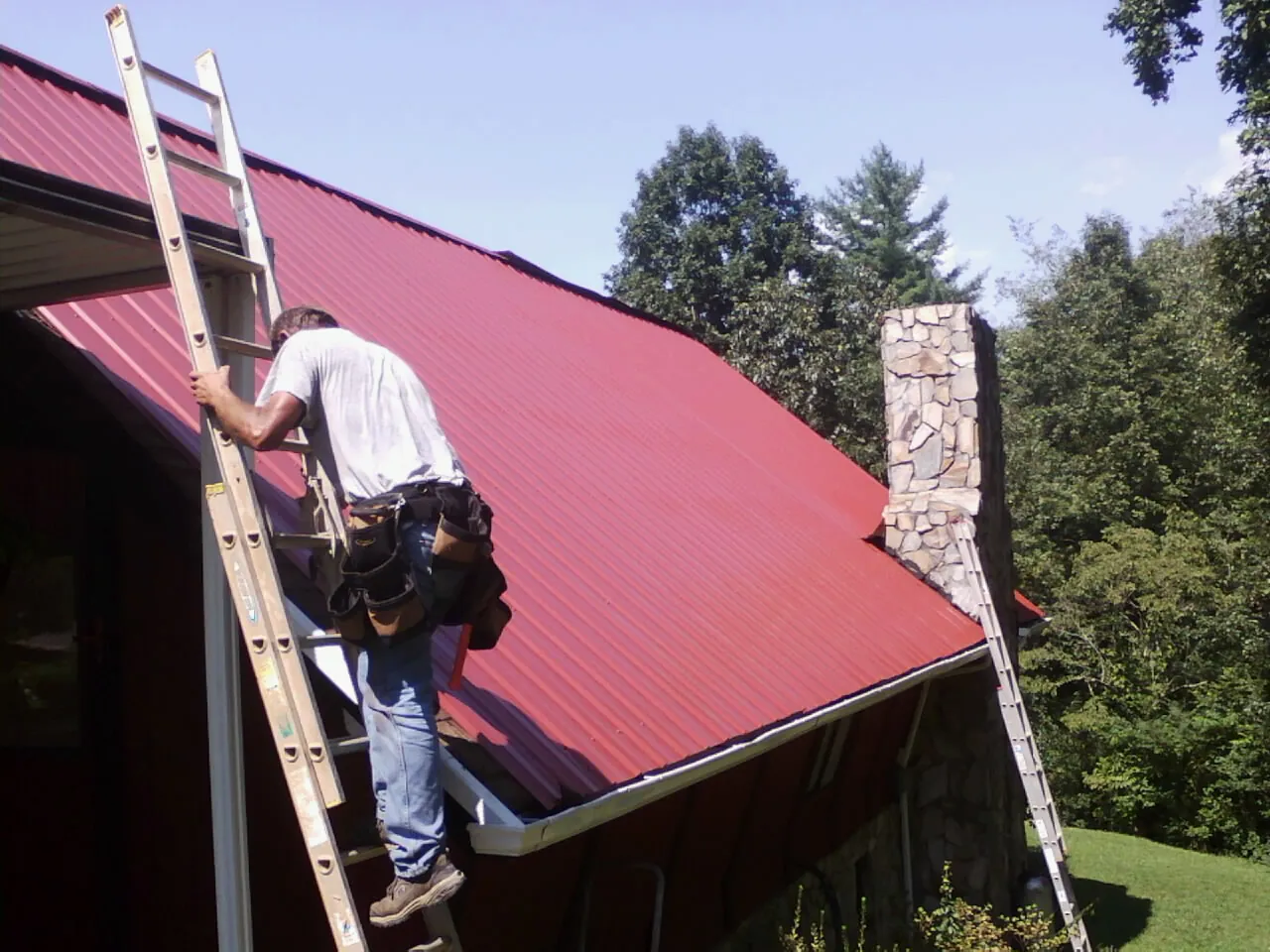 Roofer on a ladder beside a red metal roof and stone chimney — Soffit & Fascia Repair by Roofing Brothers in Oketo
